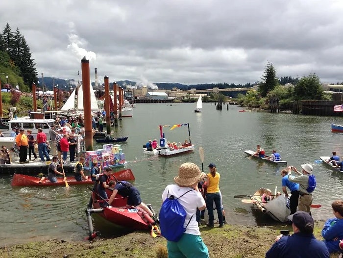 A festive boat gathering with people engaging in aquatic activities, surrounded by greenery and industrial backdrop.