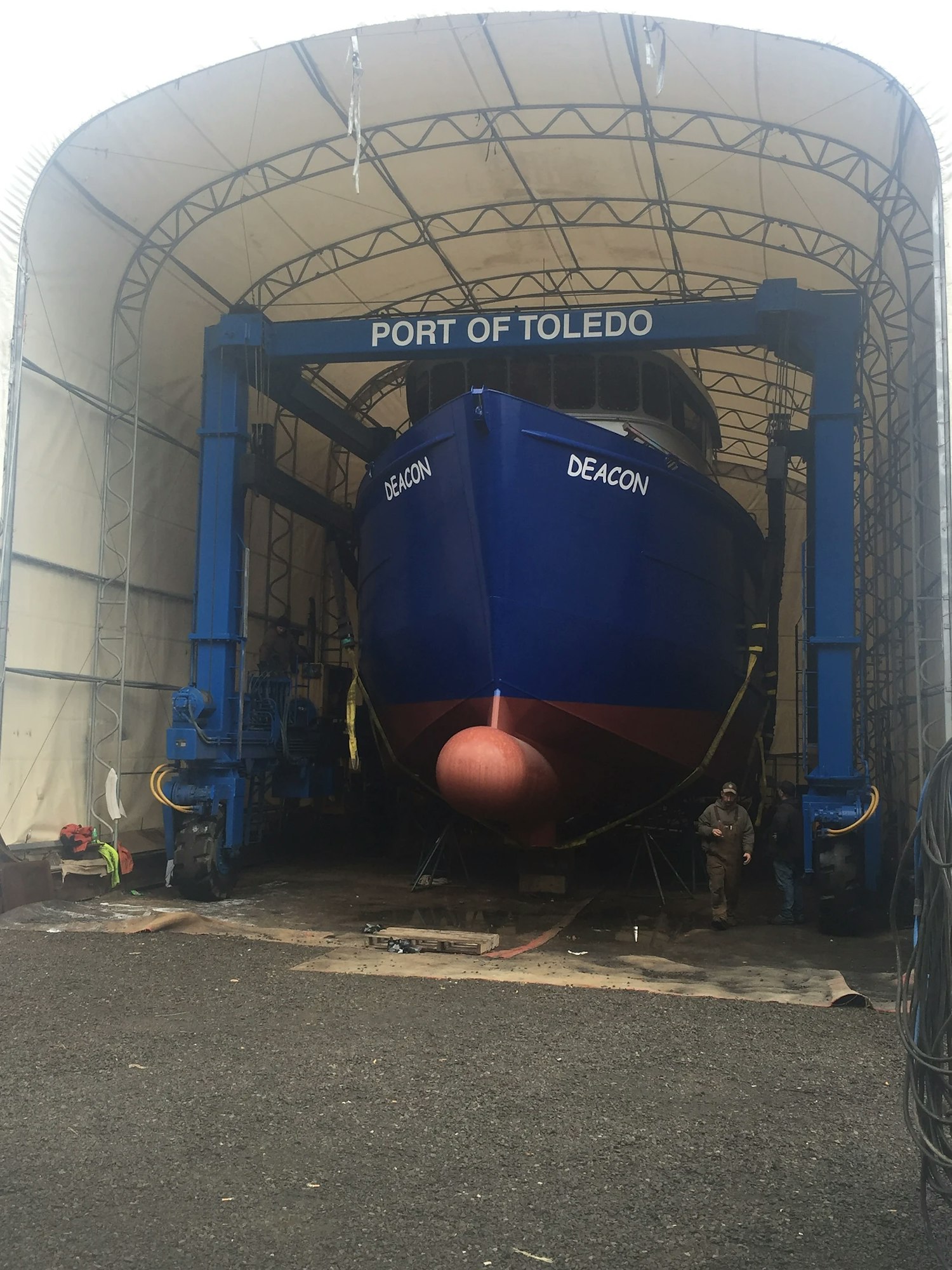 A large blue ship in a dry dock under a shelter with workers nearby.