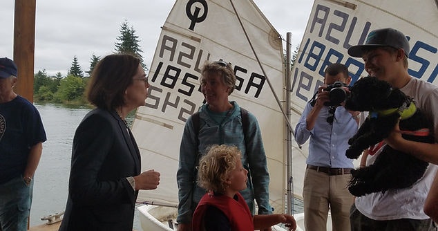 People conversing near a sail with numbers, a cameraman recording, and a person holding a dog.