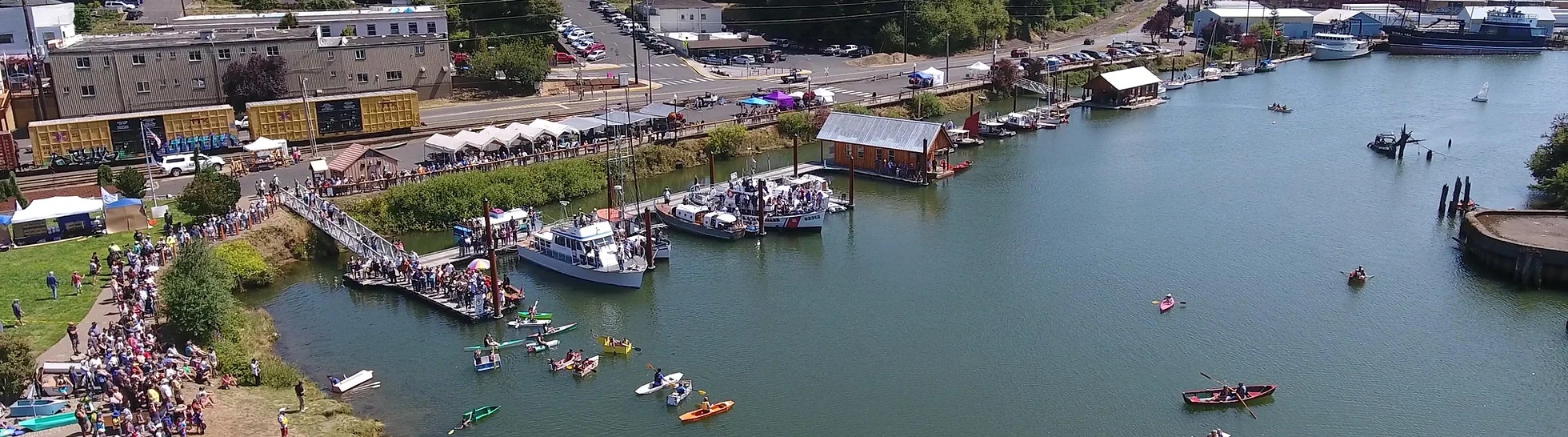 Aerial view of a waterfront with boats, crowds, floating homes, and kayakers on a sunny day.