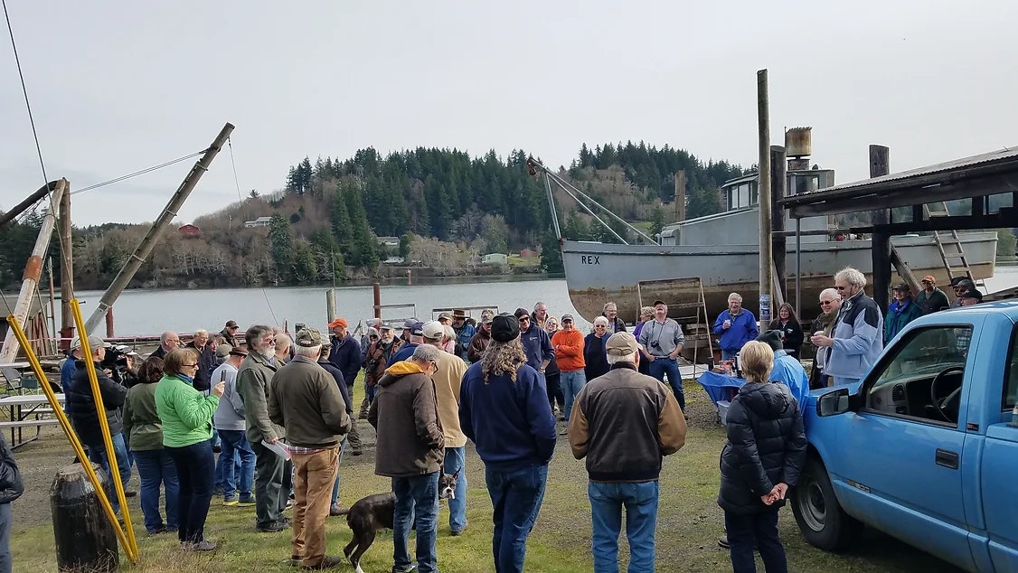 A group of people outdoors by a river with a boat and rustic equipment, possibly at a community or work event.