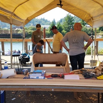 Three people at a workbench with tools, under a tent, by a waterfront.