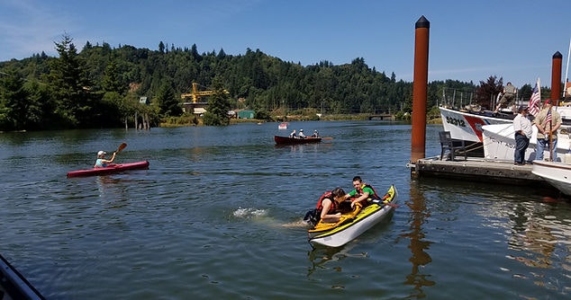 A sunny day on a river with a jet ski, kayakers, a boat, wooded hills in the background, and people on a dock.