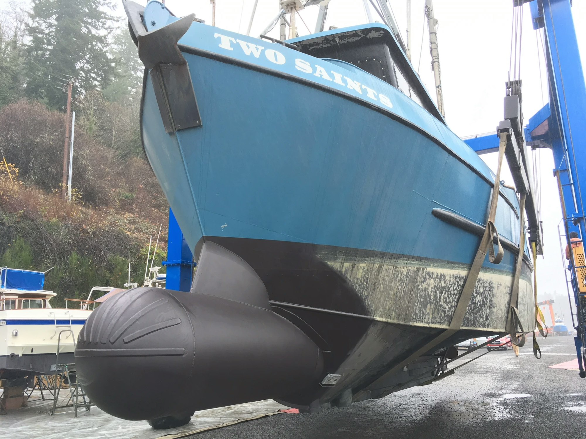 A blue boat named "TWO SAINTS" on a lift with large black fenders, dry docked for maintenance or repair.