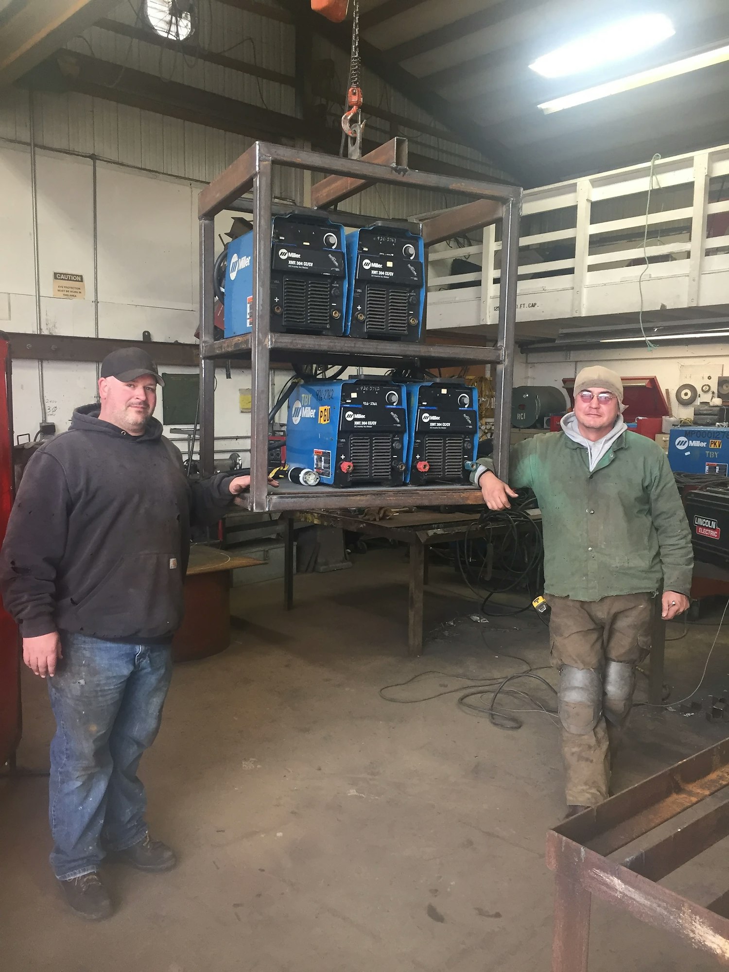 Two men in a workshop with welding equipment on a metal shelf.