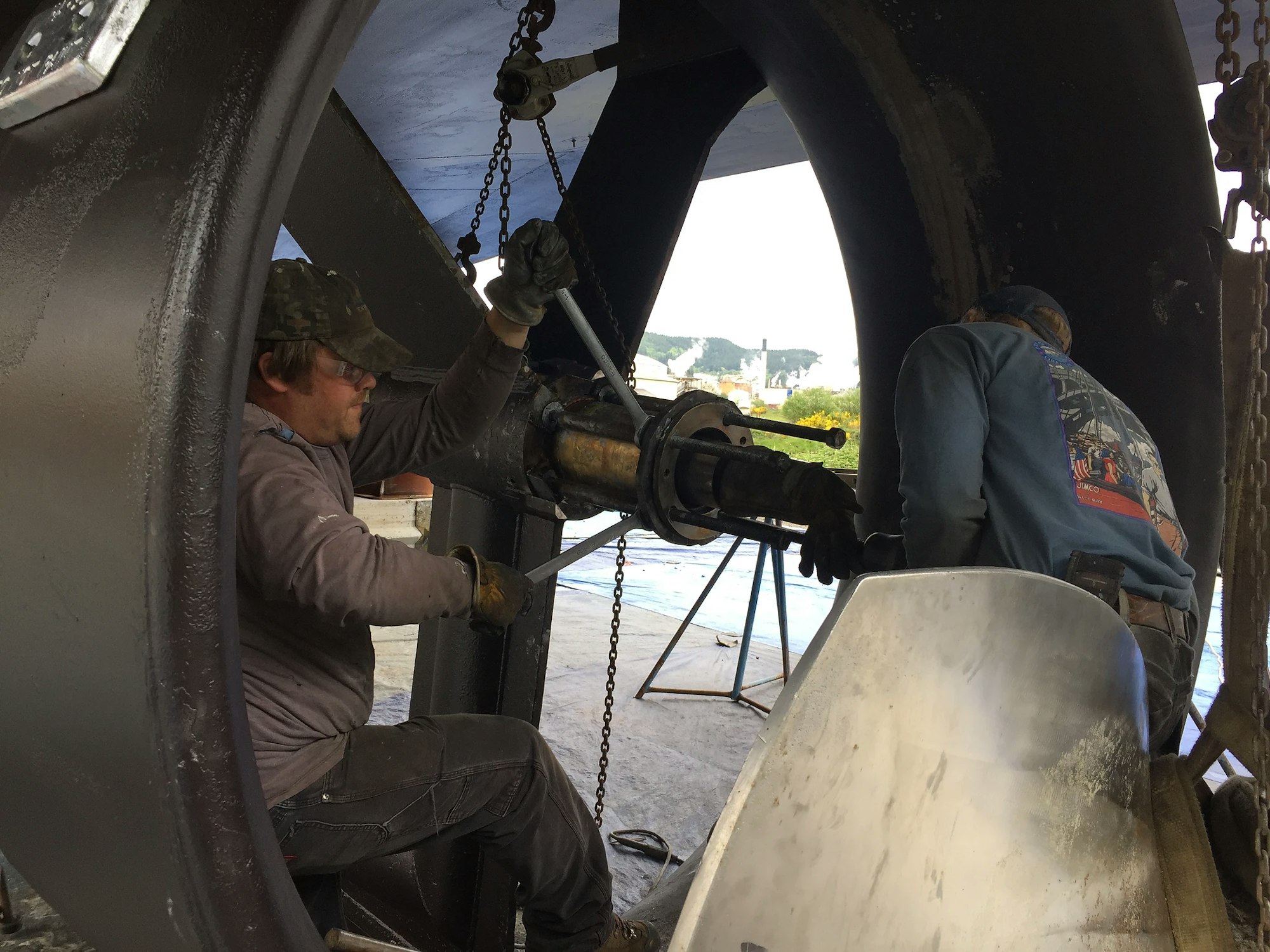 Two people repairing large machinery, likely ship or industrial equipment, in a workshop setting.