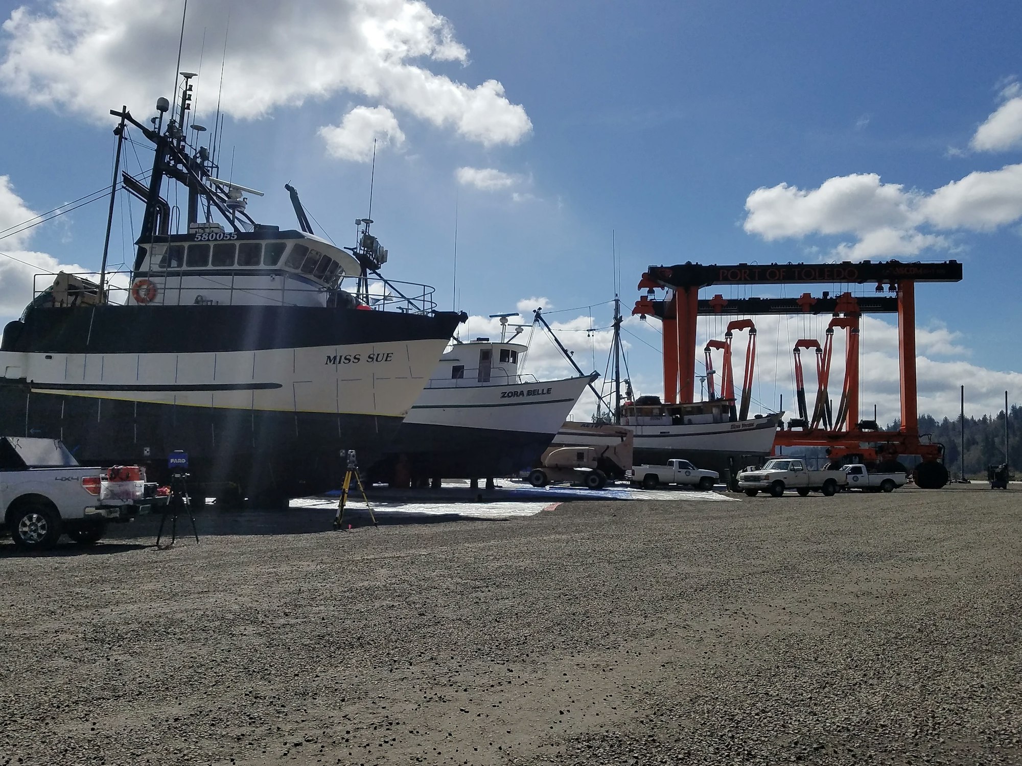 A fishing boat named "Miss Sue" on land, travel lift with a boat, vehicles, at "Port of Toledo" sign under a blue sky with clouds.