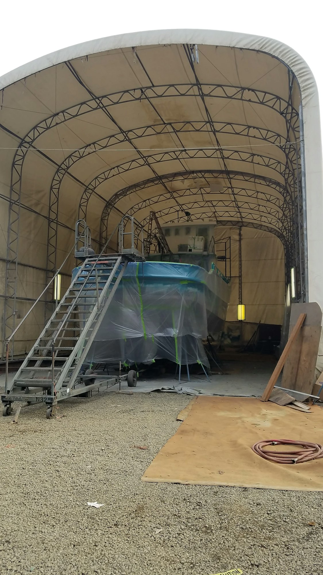 A boat wrapped in protective plastic inside a large semi-open tent with scaffolding and stairs, likely for maintenance or repair.