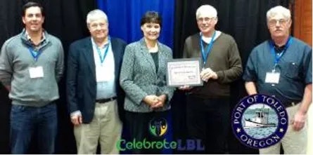 Four individuals stand behind a banner with one holding a certificate; logos indicate "Port of Toledo Oregon" and "CelebrateLBL."