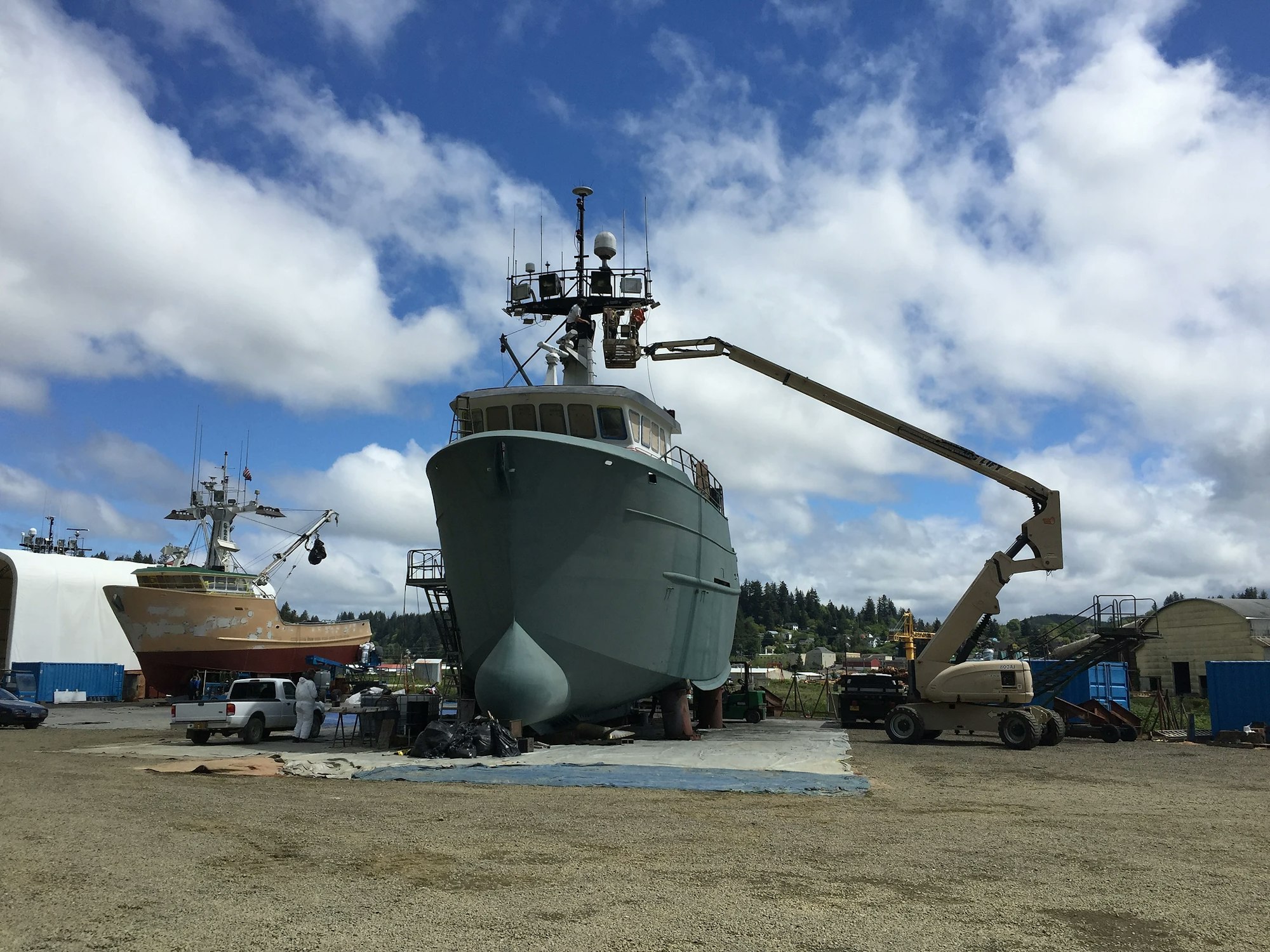 Shipyard with two large boats and a cherry picker against a blue sky with clouds.