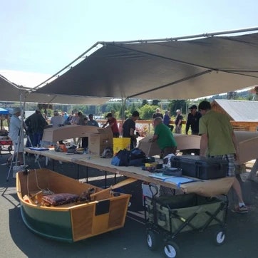 Outdoor event under tents with people, tables, a canoe, and various items on display.
