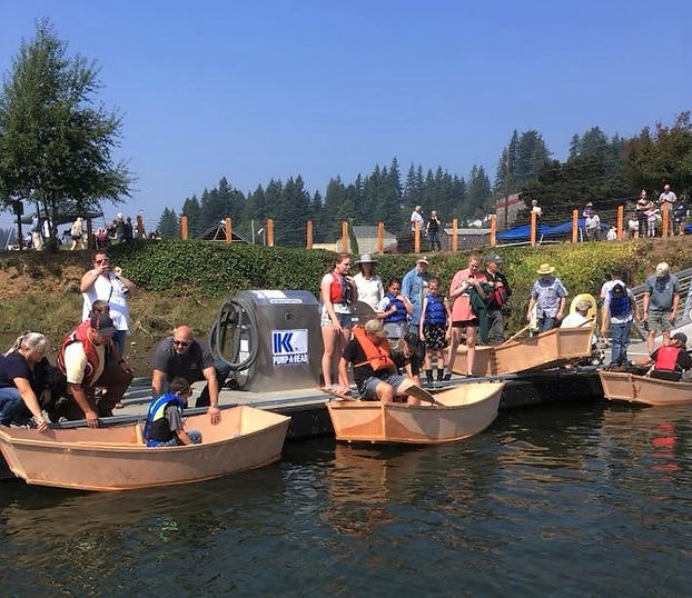 People on wooden boats preparing to row, with bystanders watching from the shore on a sunny day.