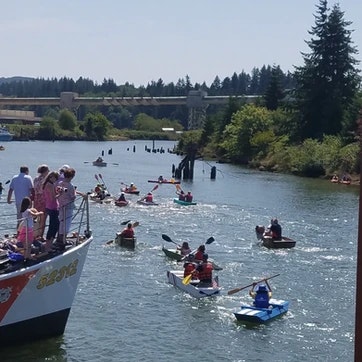 People on a dock watching kayakers on a river on a sunny day, with trees and a bridge in the background.