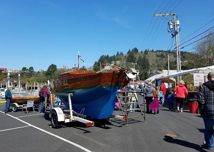 Outdoor market with a wooden boat on a trailer, people browsing, and tents with goods under a clear blue sky.