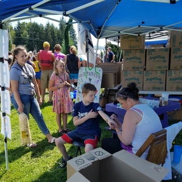 Outdoor event with people, tent, boxes, and table. Some folks stand, others engaged in an activity, possibly a community or charity event.