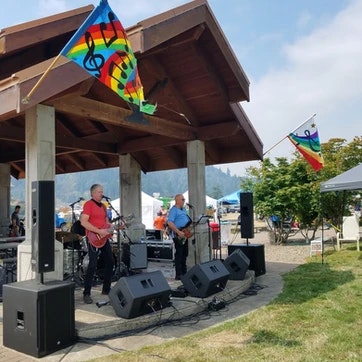 Two musicians performing outdoors, colorful flags flying, and speakers in front.