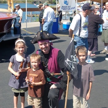 A person in pirate attire posing with three children, a boat in the background, and onlookers.