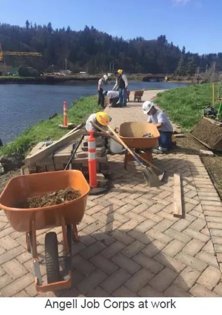 Workers in hard hats and safety vests laying bricks by a waterside.