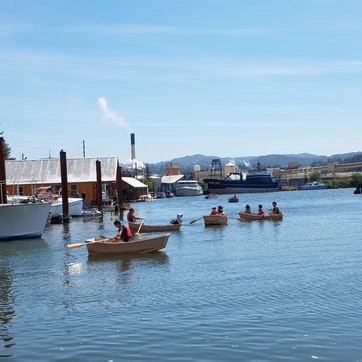 People rowing boats on a calm river with industrial buildings in the background on a sunny day.