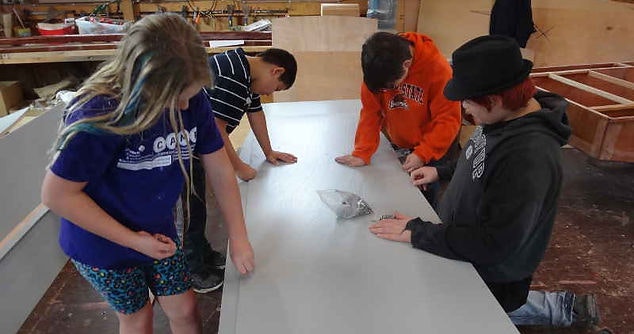 Four kids around a table, one using a hammer, crafting or learning in a workshop setting.