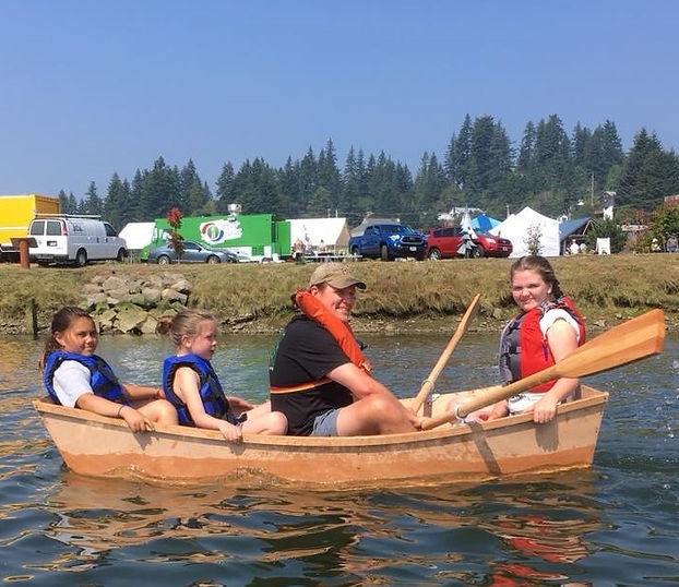 Four people in a rowboat, life jackets on, near a festival with vehicles and tents in the background.
