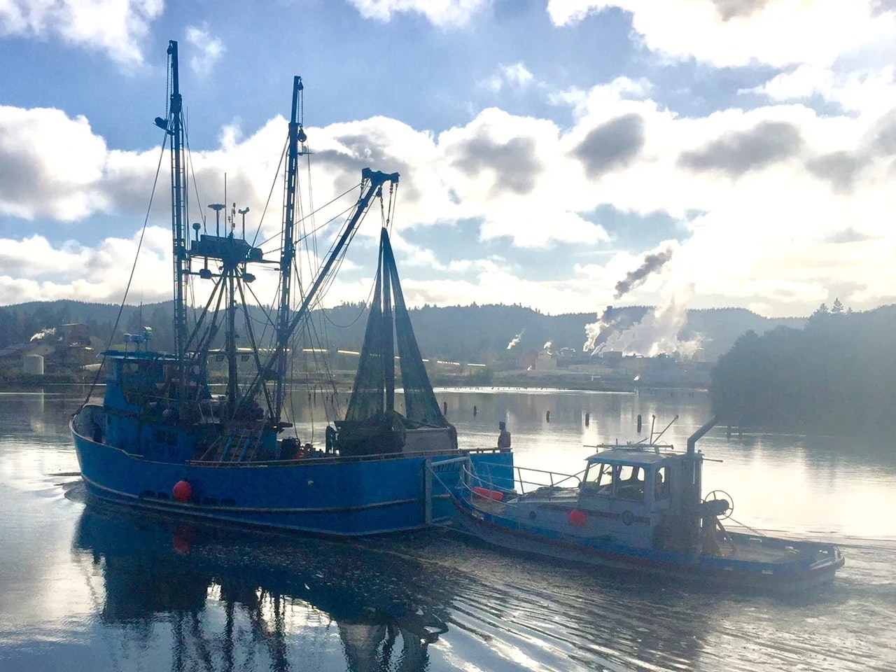 Two boats on a tranquil river with a backdrop of smokestacks and a cloudy sky.