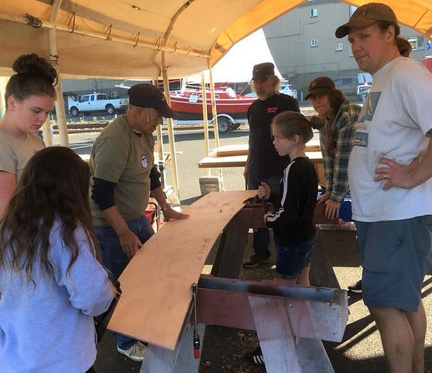 Group of people around a wooden plank at an outdoor workshop with a canopy overhead.
