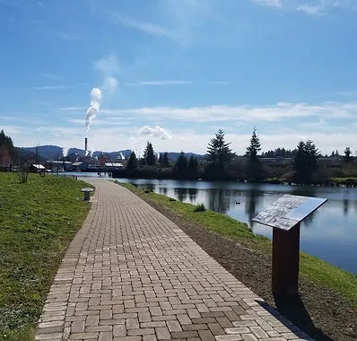 A brick pathway by a lake with trees, a smokestack in the distance, and an information board.