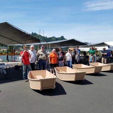 People standing near cardboard boats outside, possibly at a boat-making event or competition.