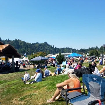 Outdoor event with people sitting on chairs and grass, blue skies, tents, and a gazebo.