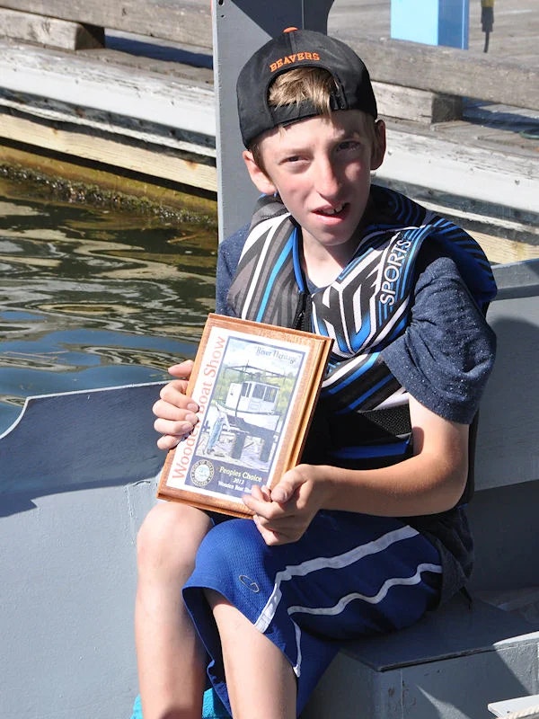 A boy in a life jacket holding a "People's Choice Award" plaque, sitting on a boat deck near water.