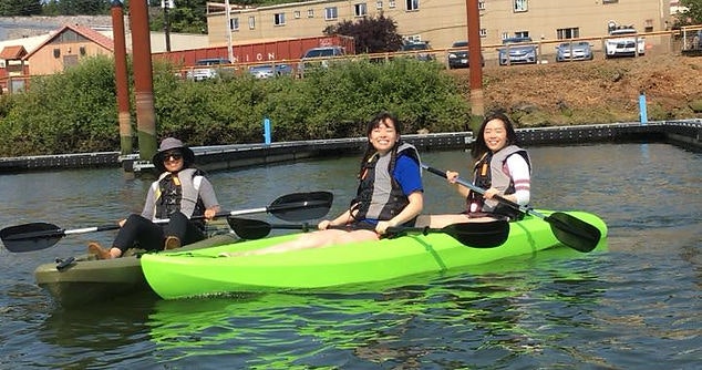 Three people in life jackets are kayaking in a green tandem kayak on a river with buildings in the background.