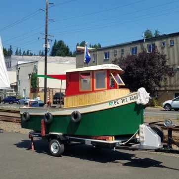 A colorful small boat on a trailer, parked on a roadside with buildings and railroad tracks in the background.