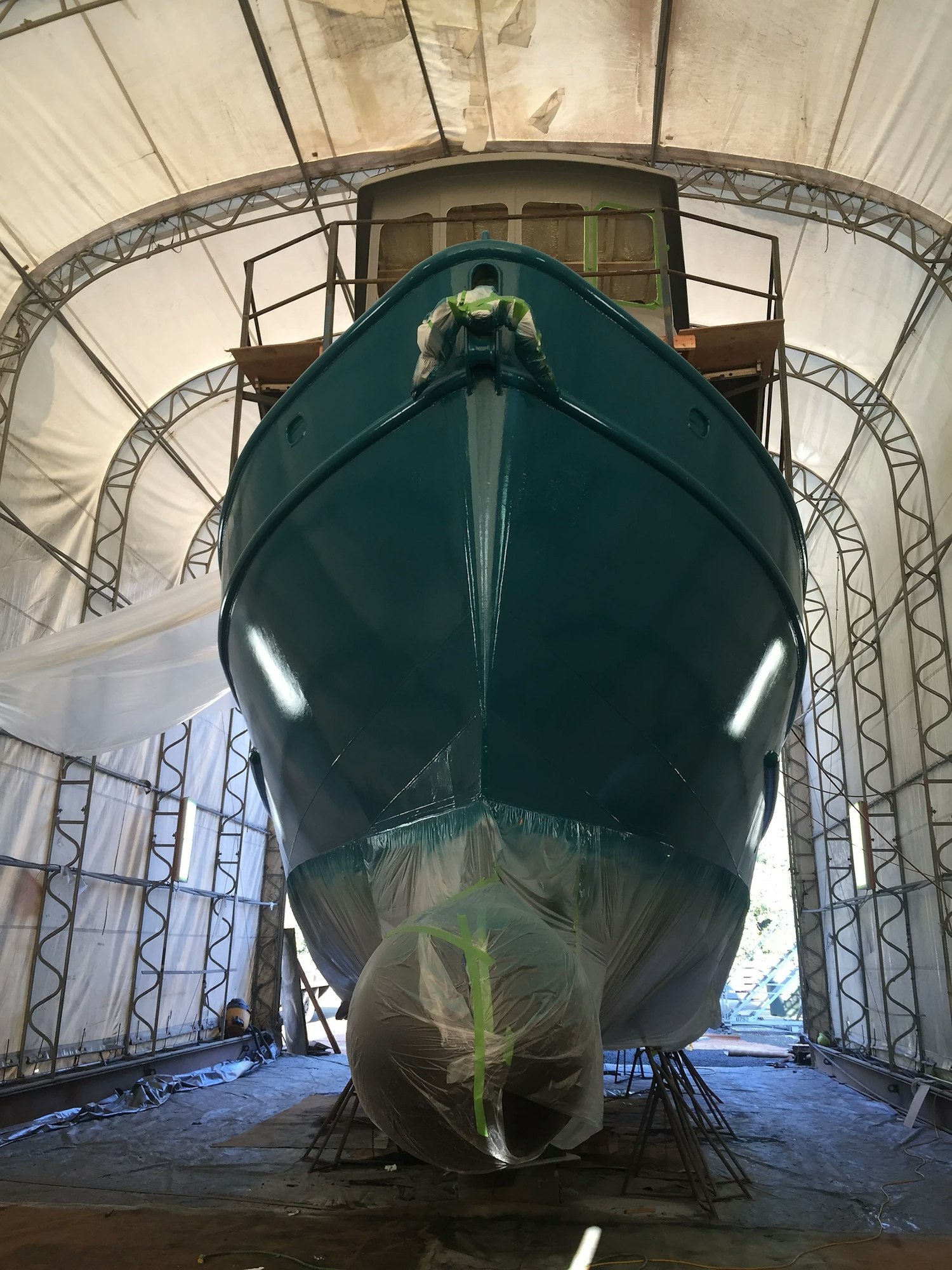 A boat in a tented area with scaffolding, likely undergoing maintenance or painting, with a person working at the bow.