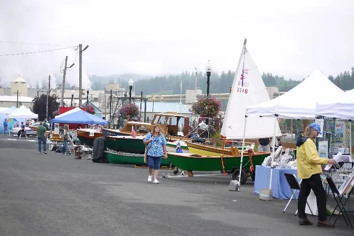 Outdoor market with stalls, colorful boats on display, and people browsing. Industrial backdrop with fog.