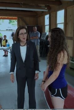Two people are talking: one in business attire and another in a swimsuit. There's an indoor setting, possibly a sports or community center.