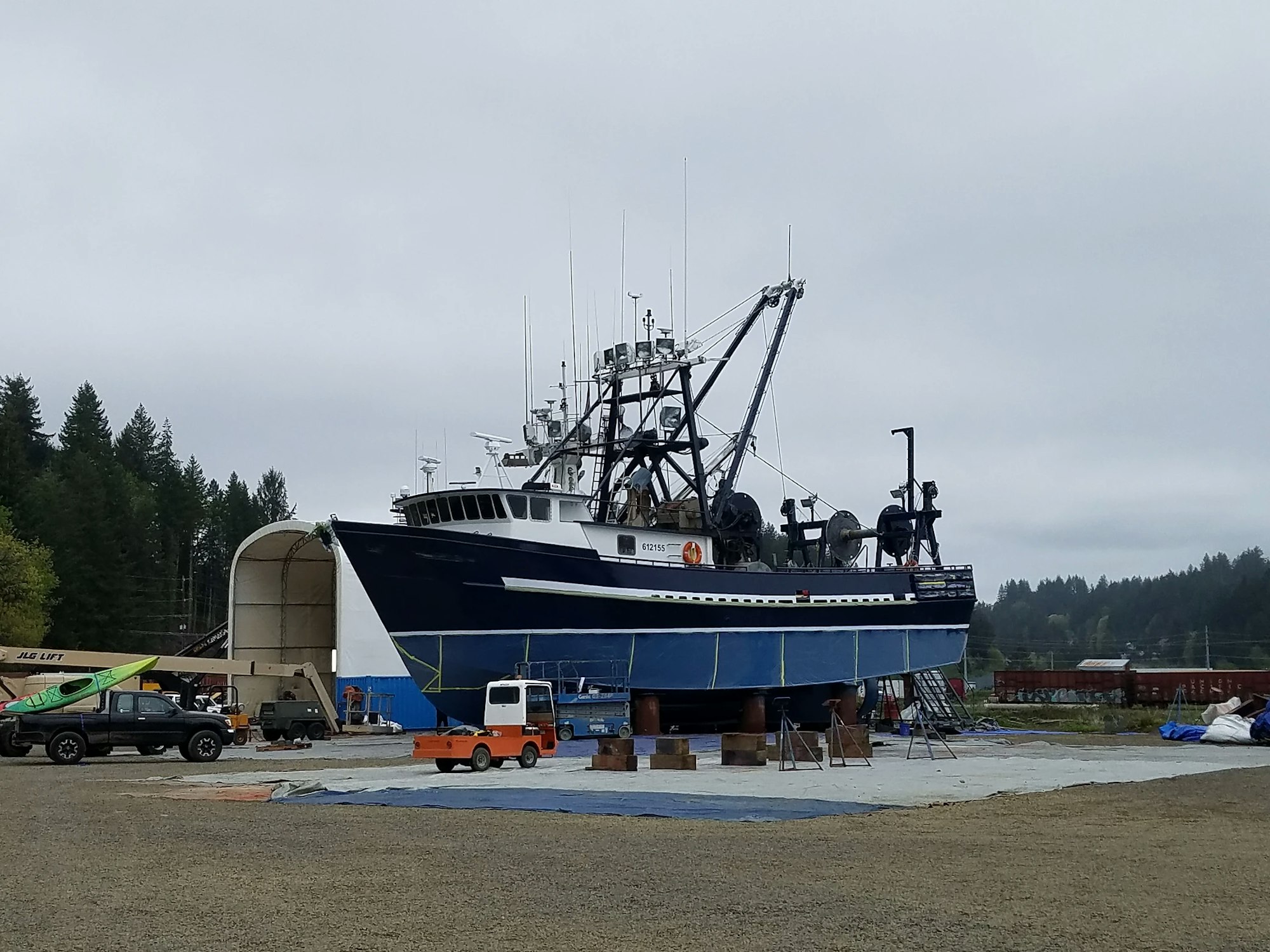A large fishing boat on dry land with forest in the background and maintenance equipment around.