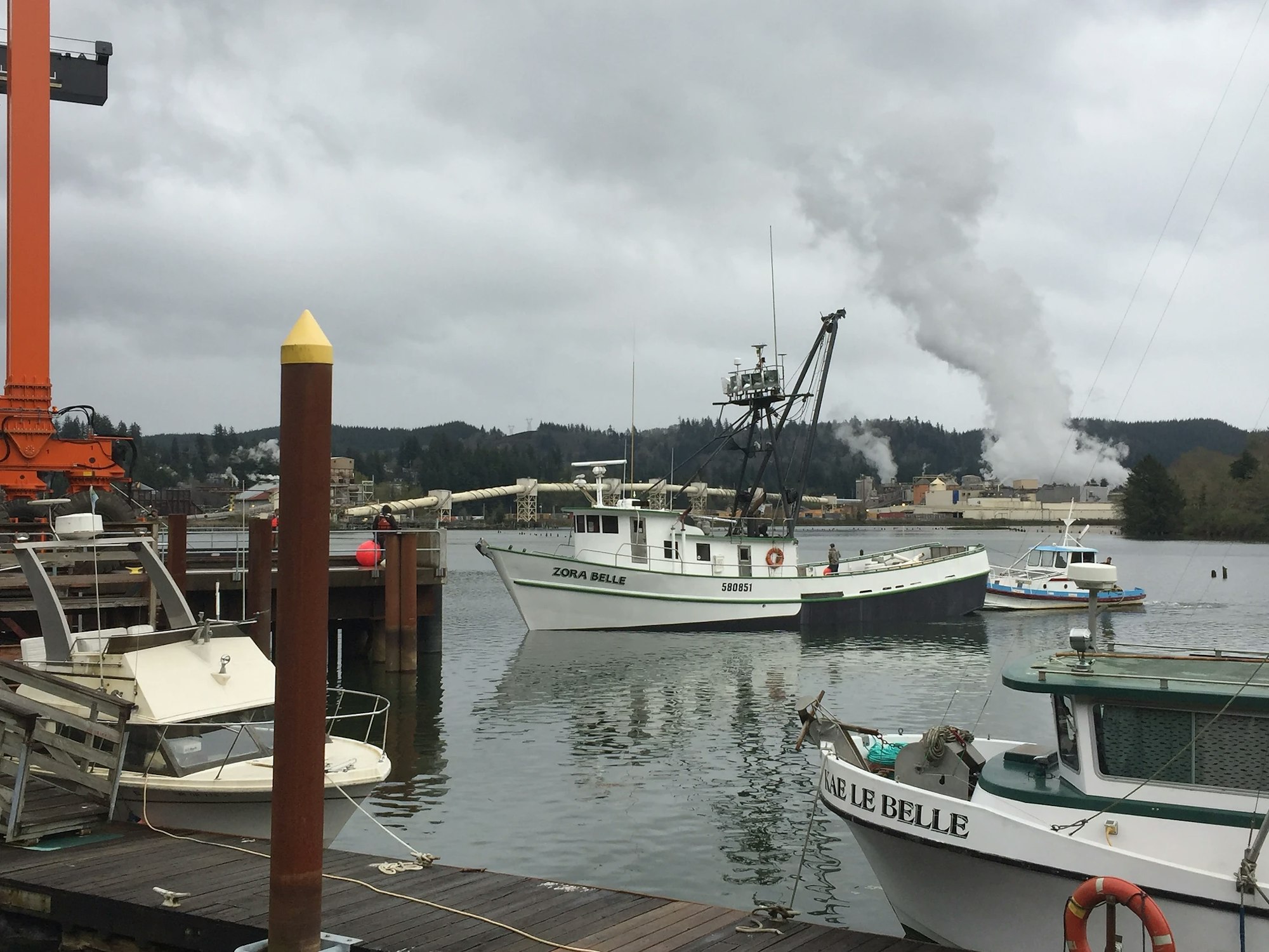 Fishing boats moored at a dock with smokestacks in the distance emitting smoke, overcast sky.