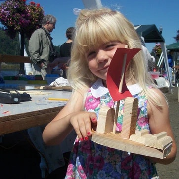 A child holding a wooden craft with a red shape on top, possibly outdoors at a market or fair.