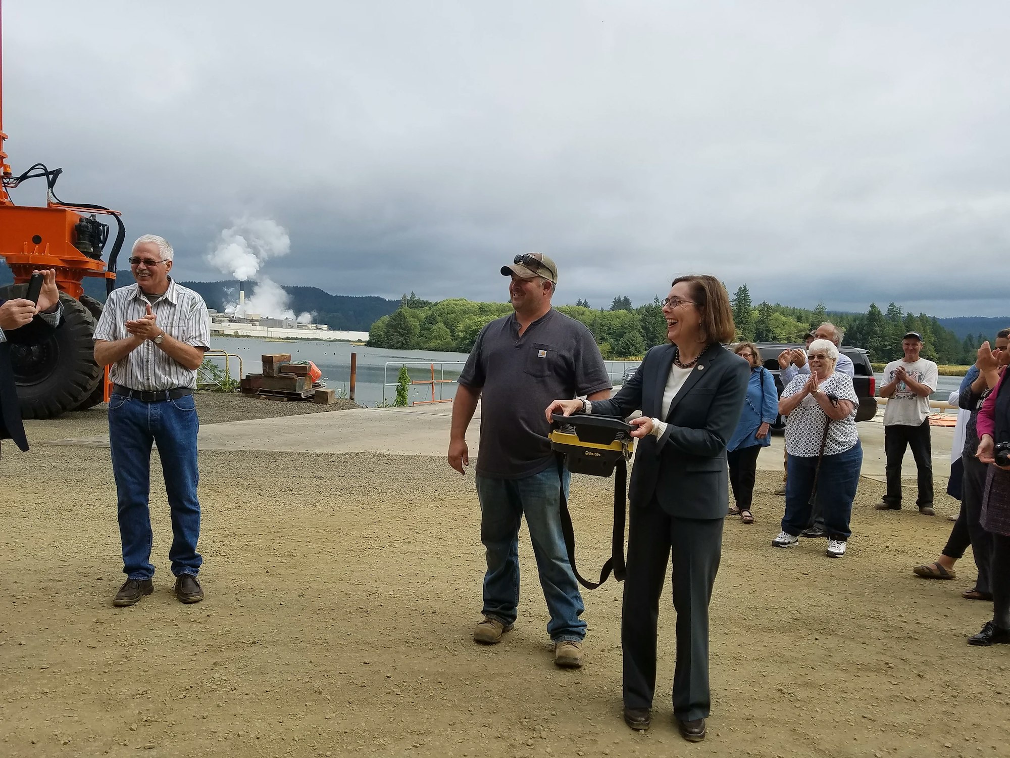Group of people outside with a woman holding a ceremonial ribbon cutting tool, industry and river in the background.