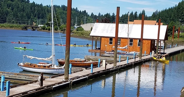 A wooden boathouse with docked boats, people kayaking on calm water, and a lush green backdrop.