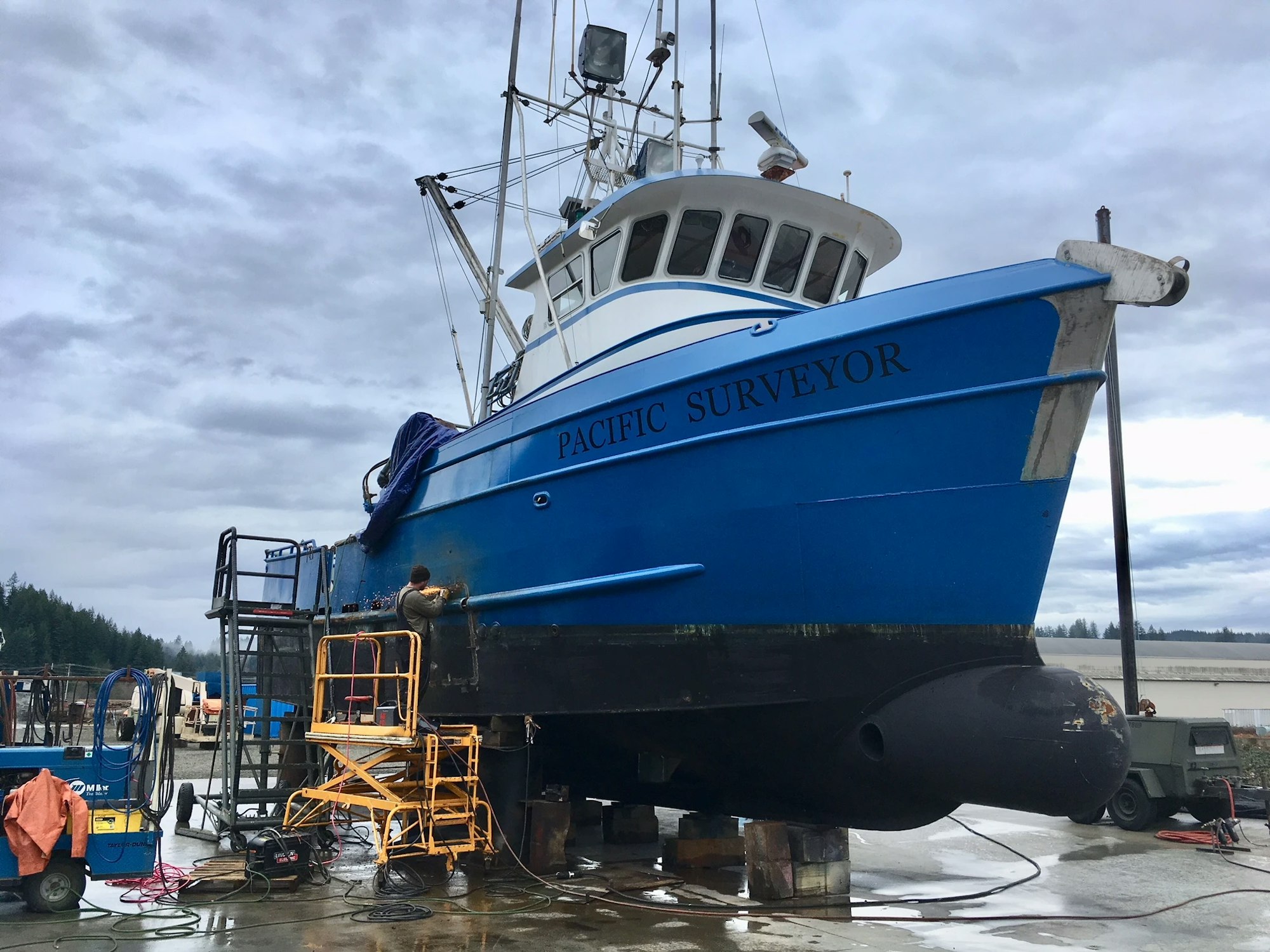 A blue boat named "PACIFIC SURVEYOR" in a dry dock with equipment around and a person working.