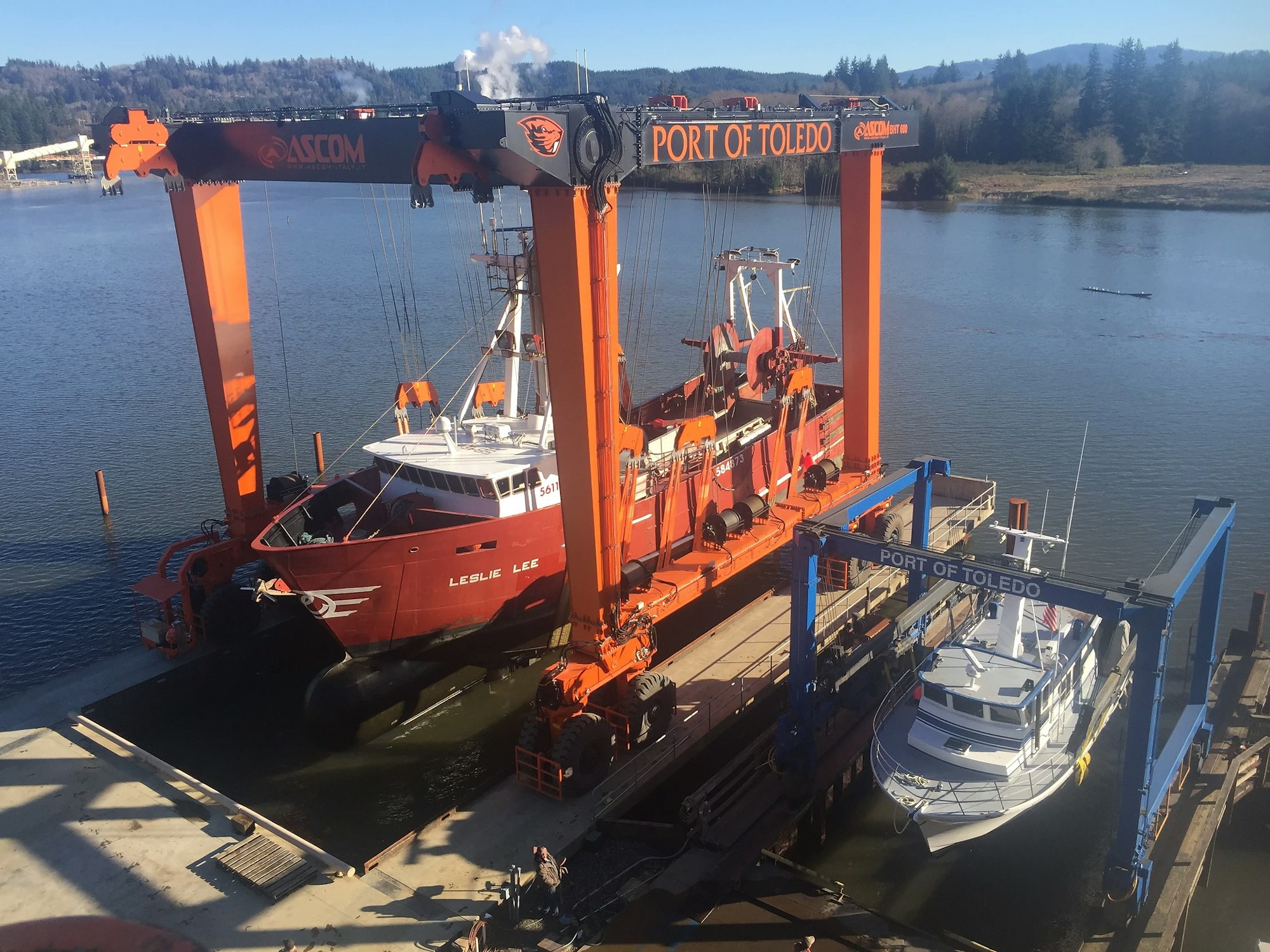 A boat lift at a port with two boats, one suspended by the lift, against a backdrop of water and hills.