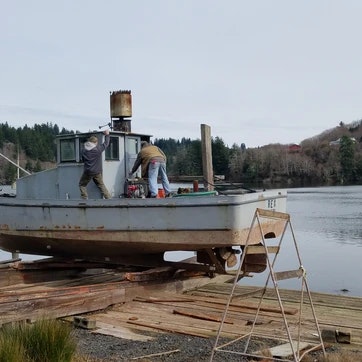 A boat on a ramp with two people working on it, a calm water body, and trees in the background.