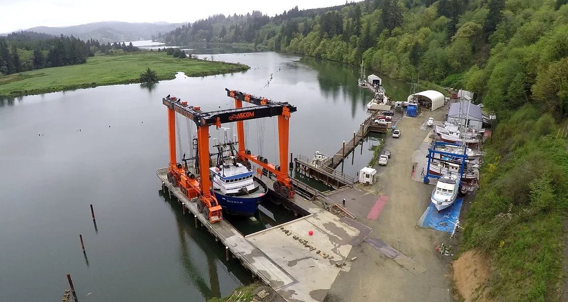 Aerial view of a shipyard with a boat lift, boats, and surrounding green landscape.