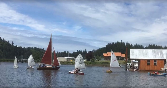Sailboats on a river with a mix of sail sizes and one with a red sail, near a boathouse and trees.