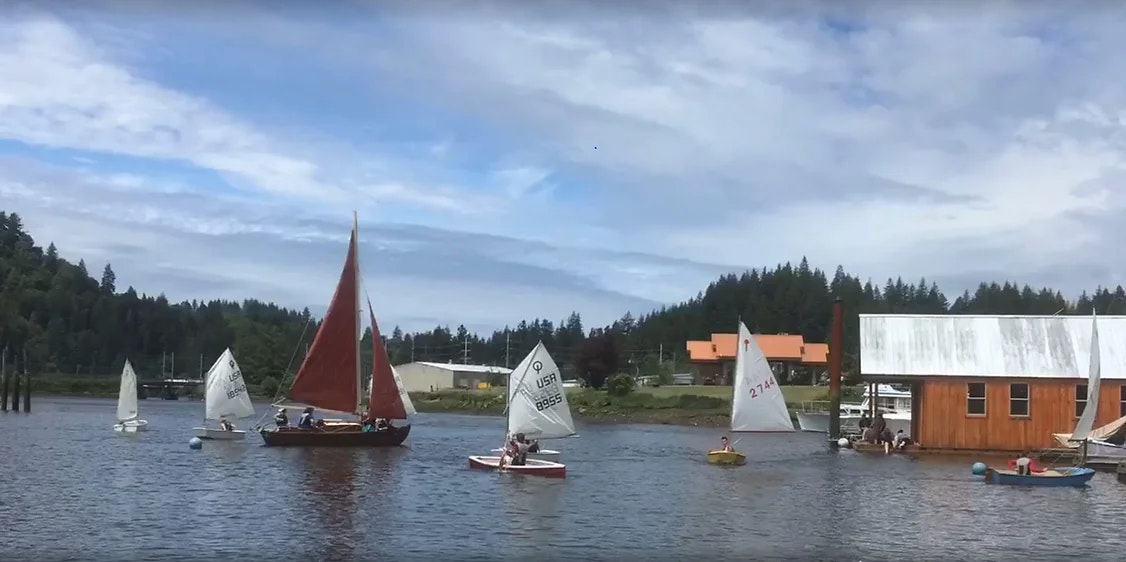 Sailing boats on water near a dock with people and a building.