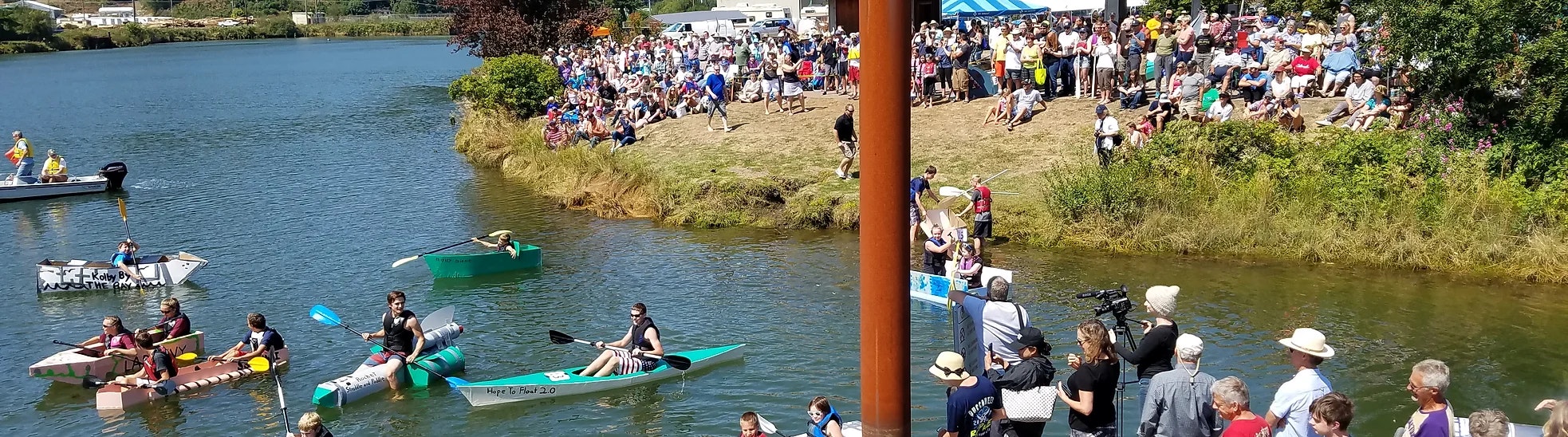 Outdoor event with people watching a cardboard boat race on a river.