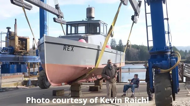 A boat named REX being hoisted by a crane, with two men standing below.
