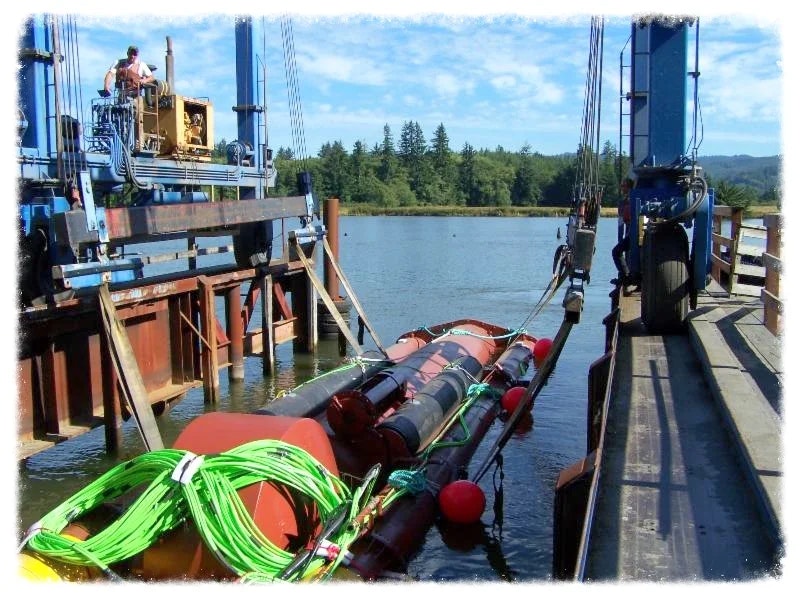 Construction equipment on a pier with cables and floating barriers on water, under a clear sky.